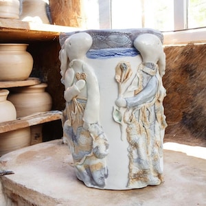 May include: A tall, white ceramic vase with relief sculptures of figures. The vase features a blue and gray rim and base, with detailed figures in flowing robes. The figures appear to be holding objects. The vase is in a pottery studio.