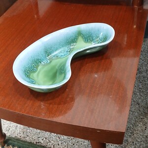 May include: A mid-century modern ceramic ashtray or dish. The ashtray is kidney-shaped with a two-tone glaze, featuring a light blue exterior and a green interior. It sits on a dark wood surface, suggesting a vintage aesthetic.