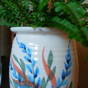 May include: A white ceramic plant pot with a floral design in blue, green, and orange. The pot is filled with green fern plants. The pot sits on a woven wicker surface.