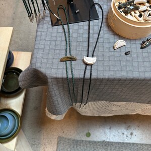 May include: Two bolo ties with different colored cords and ceramic pendants. The bolo tie on the left has a green cord and a gold crescent moon pendant. The bolo tie on the right has a black cord and a white crescent moon pendant.