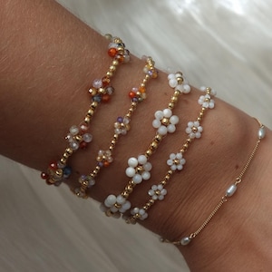 May include: A collection of beaded bracelets featuring floral and colorful designs. The bracelets are gold-toned with small, colorful beads and white flower accents. A delicate gold chain bracelet with small pearls is also visible.