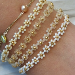 Może przedstawiać: Cztery bransoletki z koralików z złotymi akcentami i białymi lub przezroczystymi koralikami w kształcie kwiatów.