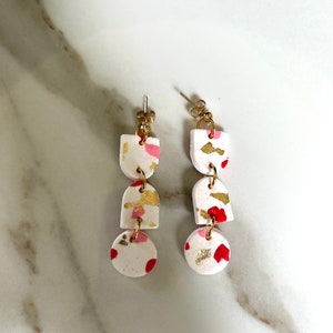 May include: A pair of dangle earrings with three white, red, and gold clay circles. The earrings have a gold hook.