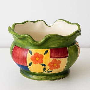 May include: A ceramic bowl with a wavy rim. The bowl is painted green, with colourful patchwork designs in red, yellow, and blue. Orange flower motifs decorate the yellow patches. The interior is off-white.