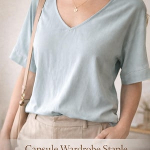 Op de afbeelding: Een lichtblauw V-hals T-shirt met korte mouwen en een beige broek. Een gouden ketting en een beige schoudertas zijn ook zichtbaar. De tekst "Capsule Wardrobe Staple" staat op de afbeelding.