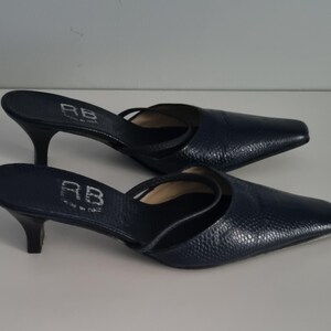 Pu&ograve; includere: Un paio di mule con tacco blu navy con punta. Le scarpe hanno una fantasia strutturata, simile alla pelle di rettile, e un cinturino sulla parte superiore del piede. I tacchi sono alti circa 6,35 cm. Le iniziali "RB" sono visibili.