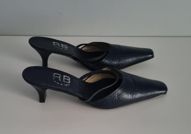 Pu&ograve; includere: Un paio di mule con tacco blu navy con punta. Le scarpe hanno una fantasia strutturata, simile alla pelle di rettile, e un cinturino sulla parte superiore del piede. I tacchi sono alti circa 6,35 cm. Le iniziali "RB" sono visibili.