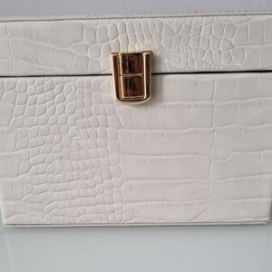 May include: A white jewellery box with a faux crocodile skin texture. The box has a rectangular shape with a hinged lid and a gold-coloured clasp. The box is designed to store jewellery and other small items.