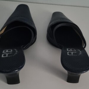 Pu&ograve; includere: Un paio di mules in pelle blu navy con punta e tacco basso. Le scarpe hanno una superficie strutturata e un cinturino sulla parte superiore del piede. I tacchi sono alti circa 5 cm.