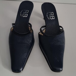 Pu&ograve; includere: Un paio di mules con tacco blu navy. Le scarpe presentano un motivo strutturato simile alla pelle di serpente e una punta affusolata. Le solette sono contrassegnate dalle lettere "RB" e "Made in Italy". Le scarpe hanno un sottile cinturino.