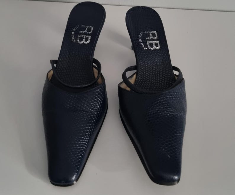 Pu&ograve; includere: Un paio di mules con tacco blu navy. Le scarpe presentano un motivo strutturato simile alla pelle di serpente e una punta affusolata. Le solette sono contrassegnate dalle lettere "RB" e "Made in Italy". Le scarpe hanno un sottile cinturino.