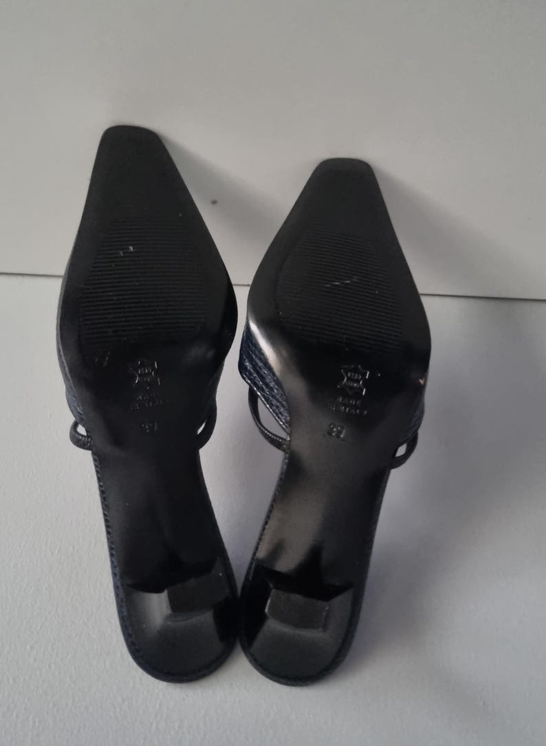 Pu&ograve; includere: Un paio di sandali con tacco blu navy. I sandali hanno la punta affilata e un sottile cinturino. Le suole sono nere con un motivo strutturato. I tacchi sono alti circa 5 cm.