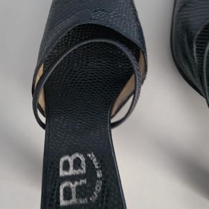 Pu&ograve; includere: Un paio di tacchi alti blu scuro a punta. Le scarpe presentano un motivo strutturato simile a quello dei rettili e sottili cinturini. I tacchi sono alti circa 7,6 cm. La suola della scarpa ha le iniziali "RB" ricamate in bianco.