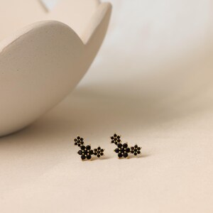 May include: Gold-tone stud earrings with a floral design. Each earring has three clusters of small, black, flower-shaped stones. The earrings are on a light surface, with a white, petal-shaped bowl in the background.