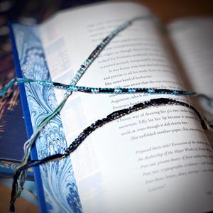 以下が含まれることがあります： 開いた本と、手作りの友情ブレスレットがいくつかあります。ブレスレットは青、緑、黒の色合いで、ビーズのアクセントが付いています。本は、大理石模様のデザインの青い表紙です。