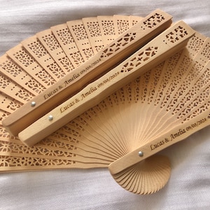 May include: Wooden hand fans with intricate cut-out designs. Two fans are closed, displaying the engraved names "Lucas & Amelia 09/08/2024". The open fan reveals a delicate, repeating pattern. The fans are a light, natural wood color.