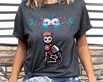 May include: A dark gray t-shirt with a black and white sugar skull cat design. The cat is wearing a flower crown and has colorful details. Two sugar skulls are above the cat with floral vines.