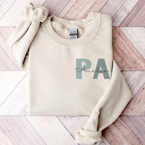 May include: A beige sweatshirt with the letters "PA" in a light green color and the name "Amelia" written in cursive underneath.