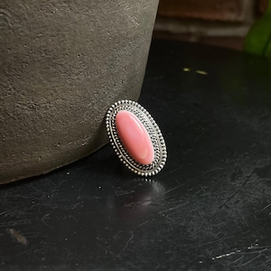 May include: Oval-shaped ring with a pink stone set in a silver-colored metal. The ring's setting has a beaded border. The ring is displayed on a dark surface, with a blurred background.