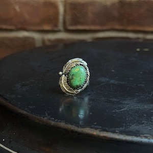 Puede incluir: Un anillo de plata con una piedra ovalada de turquesa verde brillante. La piedra está enmarcada por un diseño de cuerda trenzada y detalles de plumas. El anillo está sobre una superficie oscura y texturizada.