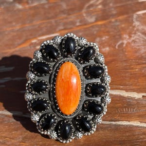 Pode incluir: Anel de prata oval com uma pedra central laranja e pedras pretas circundantes. O anel tem um design detalhado, semelhante a uma flor, em torno das pedras pretas. O anel está sobre uma superfície de madeira.