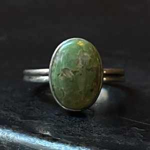 Puede incluir: Un anillo de plata con una piedra ovalada de color verde claro. La piedra tiene un aspecto moteado con inclusiones de color verde oscuro y marrón. La banda del anillo es de diseño simple de doble banda.