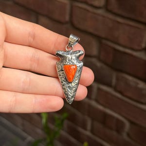 May include: A silver-toned pendant in the shape of an arrowhead, featuring a vibrant orange stone at its center. The pendant has a textured surface and a loop for a chain. The background is a brick wall.