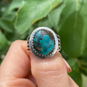 Puede incluir: Un anillo de plata con una gran piedra turquesa de forma ovalada con inclusiones de matriz de color marrón oscuro. La piedra está engastada en un bisel dentado y la banda del anillo consta de varios hilos de plata paralelos.