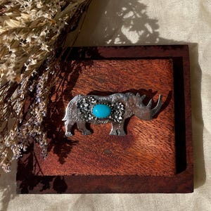Pode incluir: Um broche de rinoceronte em tom prateado com uma pedra turquesa oval central rodeada por pequenos detalhes metálicos florais. A figura do rinoceronte apresenta uma superfície texturizada e um chifre proeminente, exibindo um design detalhado.
