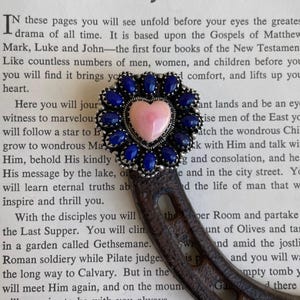 May include: A decorative object featuring a dark metal horseshoe base topped with a circular cluster of oval blue stones surrounding a central light pink heart-shaped stone. The piece is positioned over a page titled The Story of Jesus.