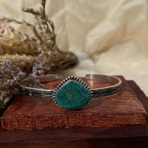 May include: A silver cuff bracelet featuring a central teardrop-shaped turquoise stone with dark matrix veining. The metal band has a textured, oxidised finish with small engraved geometric patterns along the sides and a beaded border around the stone.