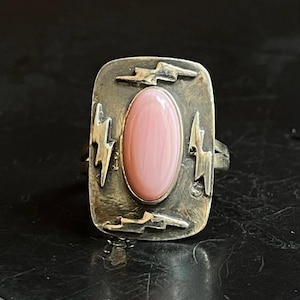 May include: A silver ring with a rectangular face featuring a light pink oval stone. The ring is adorned with four lightning bolt designs around the stone. The ring has a vintage or antique appearance.