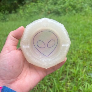 May include: A white, iridescent, geometric ashtray with an alien face design. The ashtray is made of resin and has a smooth, glossy finish.