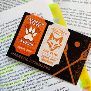 Op de afbeelding: Twee magnetische bladwijzers met een foxhole court en palmetto state thema. De bladwijzers zijn oranje en wit met een vos en een pootafdruk ontwerp. De tekst op de bladwijzers luidt "Foxhole Court", "Exy Game", "Palmetto State", "Foxes", "Admit One", en "Magnetic Bookmarks".