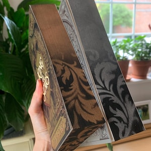 May include: Two hardcover books with decorative covers. One book has a brown cover with gold lettering and a floral design. The other book has a black cover with a silver floral design. The books are held at an angle, showcasing the side edges.