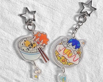 May include: Two anime-style keychains with star-shaped clasps. One keychain features a bowl of rice with a cartoon character using chopsticks. The other keychain shows a plate of food with a cartoon character.