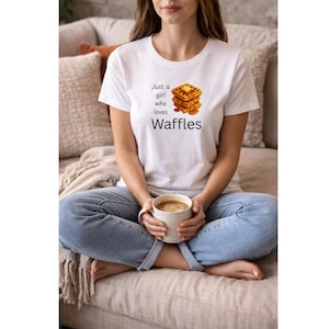 Könnte beinhalten: Weißes T-Shirt mit dem Text "Just a girl who loves Waffles" und einer Waffel-Grafik. Die Person trägt eine blaue Jeans und hält eine Tasse Kaffee. Ein lässiges Kleidungsstück für den Alltag.