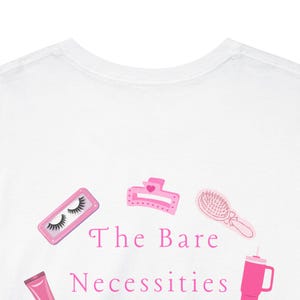 May include: White t-shirt with a pink graphic design featuring beauty essentials. The design includes illustrations of false eyelashes, a hair clip, a hairbrush, and a tumbler. The text "The Bare Necessities" is also included.