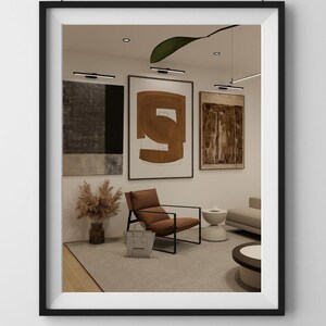 May include: Interior design with framed abstract art in neutral tones. A brown leather armchair with a black frame is positioned in front of the artwork. A grey handbag rests on the floor. The room has a modern, minimalist aesthetic.