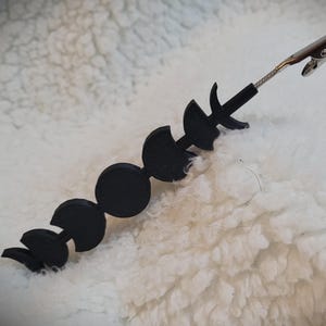 May include: A black metal clip with a series of crescent moon shapes attached. The clip is silver and has a spring mechanism.