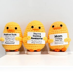 May include: Three yellow crocheted duck figurines holding signs with positive messages. The signs read "Positive Duck", "You're Ducking Awesome", and "Mom I Ducking Love You!" Each sign includes the phrase "Tiny duck is here to always remind you!"