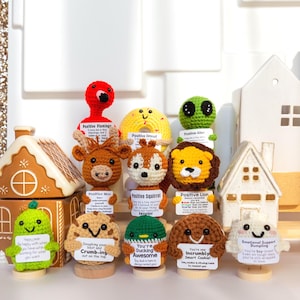 May include: A collection of crocheted animal figurines, including a flamingo, donut, alien, deer, squirrel, lion, and others. Each figure holds a sign with positive affirmations. The figurines are displayed with decorative gingerbread houses and a white house figurine.