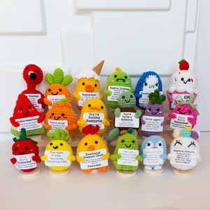May include: A collection of colorful, handmade crochet figures, including a flamingo, watermelon, orange, duck, dinosaur, and penguin. Each figure holds a small sign with positive messages. The figures are displayed on small wooden stands.