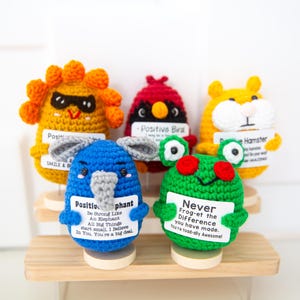 May include: A collection of five colorful, crocheted animal figurines: a lion, a bird, a hamster, an elephant, and a frog. Each figure holds a small sign with positive messages. The figurines are displayed on a wooden shelf.