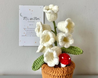 May include: A crocheted flower arrangement featuring white bell-shaped flowers, green leaves, and a red mushroom in a brown pot. A small card with the text "Moon Fam" is in the background. The arrangement is handmade.