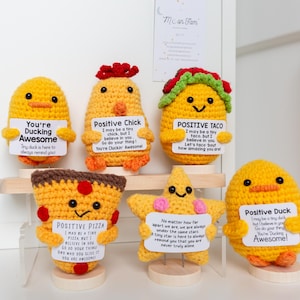 May include: A collection of handmade crochet figures, including a duck, chick, taco, pizza, and star, each holding a sign with positive messages. The figures are yellow, orange, and brown, with black eyes and various decorative elements.