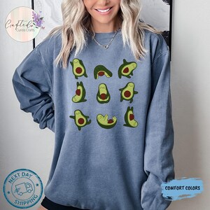 May include: A blue sweatshirt featuring a pattern of green avocado illustrations in various yoga poses. The avocados have brown seeds and are set against a blue background. The sweatshirt has a crew neck and long sleeves.