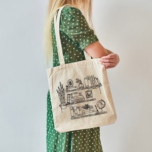 May include: A white canvas tote bag with a black line drawing of a bookshelf with books, plants, and a globe. The bag has a long strap and is being held by a person wearing a green polka dot dress.