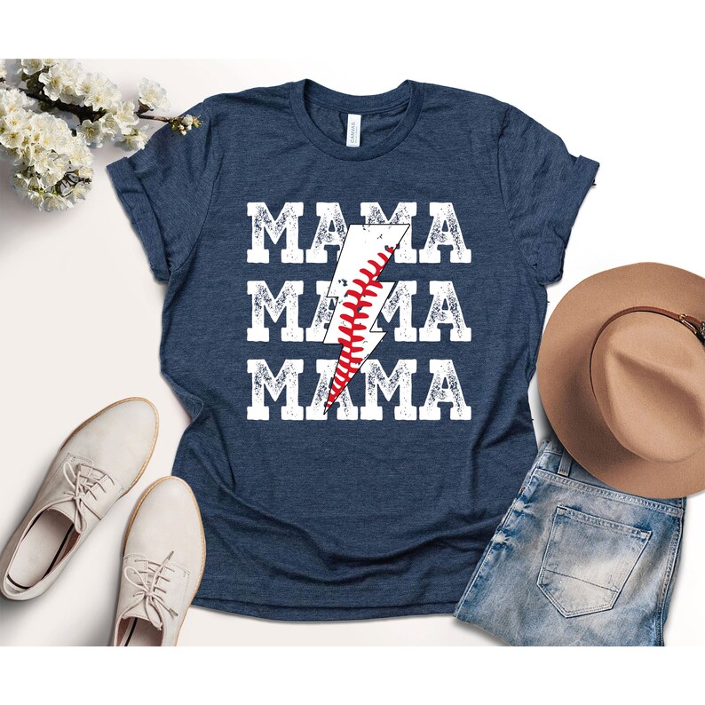 以下が含まれることがあります： 紺色のTシャツに、くたびれた白のグラフィックで「MAMA」という文字が3回繰り返されています。グラフィックの中央には、野球の縫い目が施された稲妻があります。