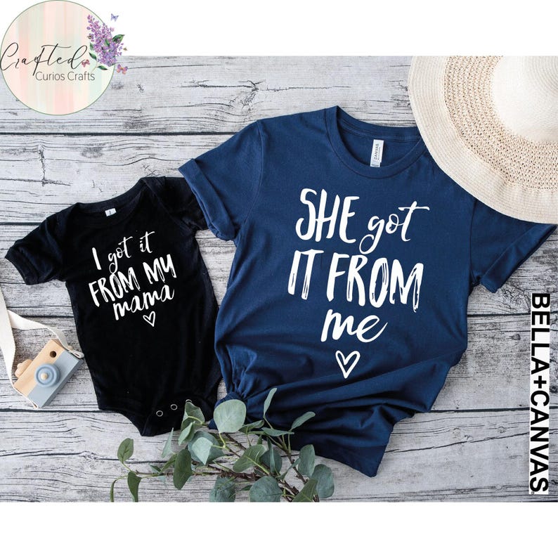 Puede incluir: Dos camisetas con un dise&ntilde;o a juego. La camiseta m&aacute;s grande es azul marino con texto blanco que dice "SHE got IT FROM me" con un coraz&oacute;n. La camiseta m&aacute;s peque&ntilde;a es negra con texto blanco que dice "I got it FROM MY mama" con un coraz&oacute;n.